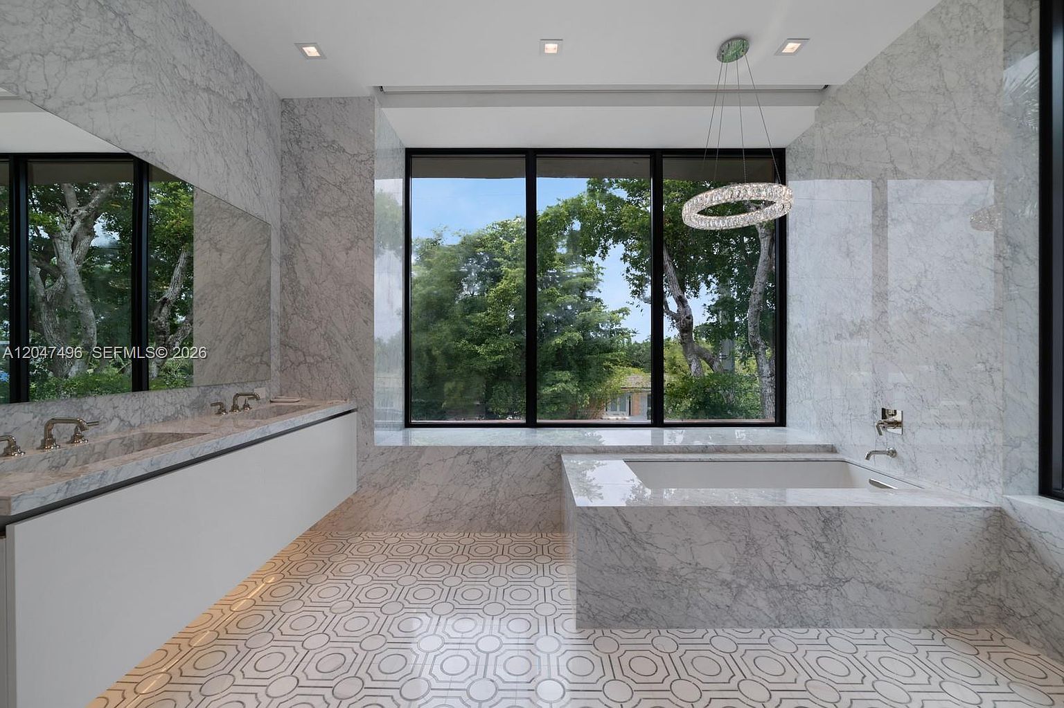 This luxurious primary bathroom features extensive stone wall cladding and a striking geometric patterned tile floor. A large soaking tub is positioned beneath expansive windows that offer a clear view of the surrounding trees, while a floating vanity with dual sinks sits opposite. The space is illuminated by a modern, circular pendant light, creating a bright and sophisticated atmosphere.