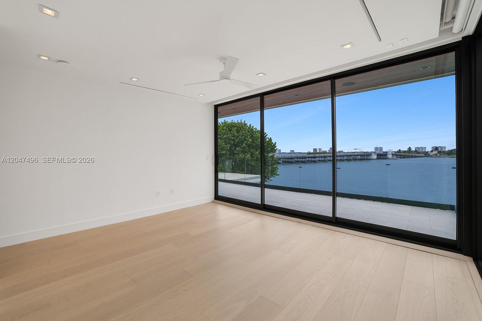 This minimalist interior space features light wood flooring and crisp white walls, creating a bright and airy atmosphere. A large sliding glass door provides a seamless transition to an outdoor balcony, offering expansive, unobstructed views of the water and distant cityscape. The room is finished with a modern ceiling fan and recessed lighting, emphasizing a clean, contemporary aesthetic.