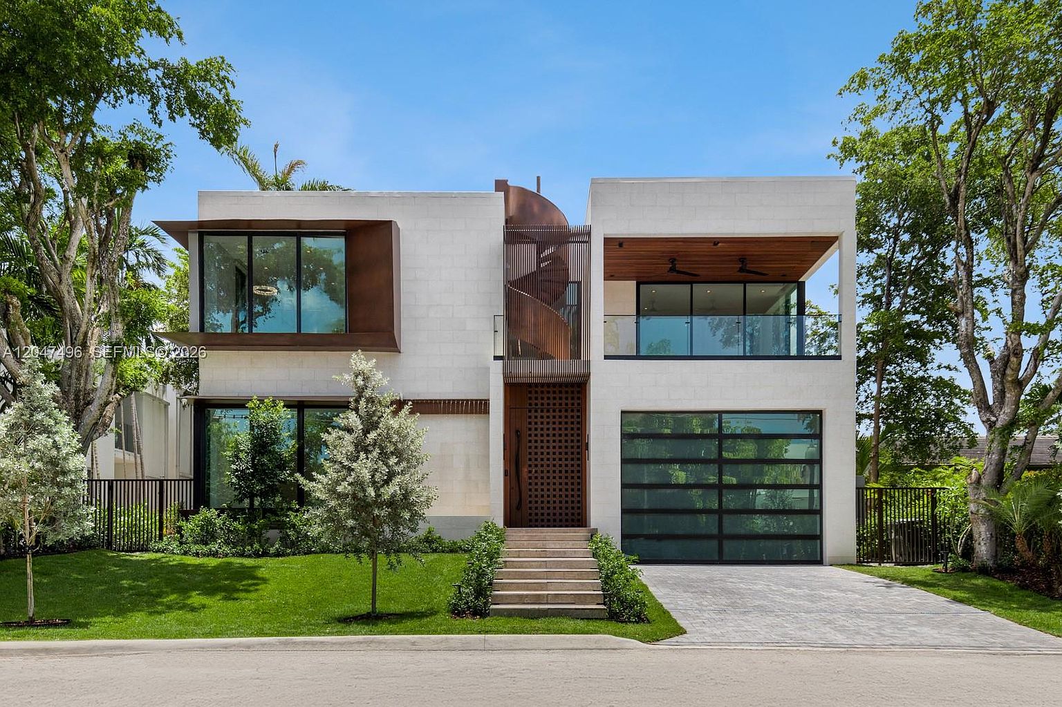 This modern two-story residence features a striking facade with a combination of light-colored stone masonry, dark metal accents, and large glass panels. The home includes a prominent spiral staircase enclosed by vertical slats, a spacious upper-level balcony with wood-paneled ceilings, and a sleek glass-paneled garage door. The property is framed by mature trees and a manicured lawn, presenting a sophisticated and contemporary architectural aesthetic.