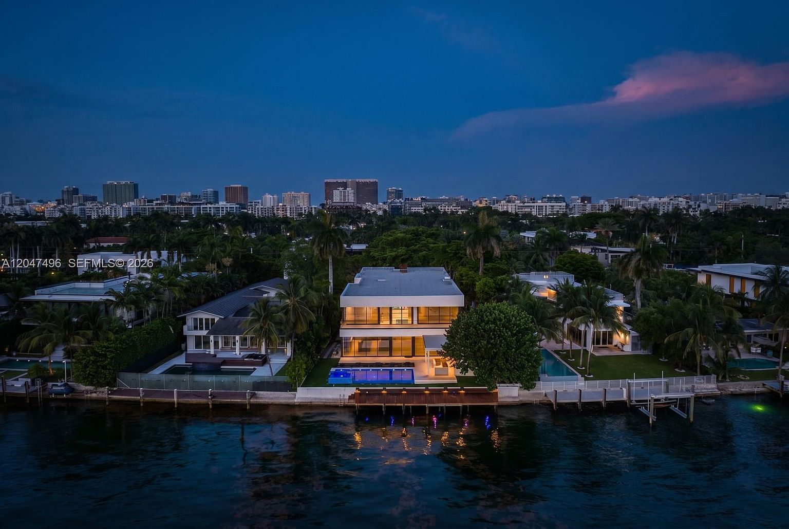 This aerial perspective captures a contemporary waterfront residence at dusk, showcasing its sleek, multi-level architectural design and expansive glass facade. The property features a private swimming pool, a wooden dock extending into the water, and lush landscaping, all set against a vibrant city skyline backdrop. The scene conveys a sense of luxury and tranquility, highlighting the seamless integration between the modern home and its coastal environment.