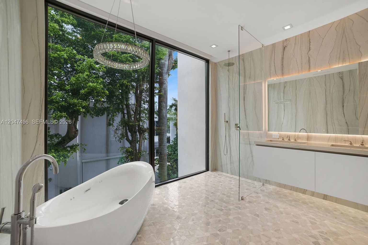 This luxurious primary bathroom features a sleek, freestanding soaking tub positioned next to expansive floor-to-ceiling windows that offer a serene view of lush greenery. The space is finished with light-toned, patterned tile flooring and a large, wall-mounted vanity with a full-width mirror and integrated lighting. A glass-enclosed walk-in shower adds to the open, airy feel, while a modern circular chandelier provides a sophisticated focal point.