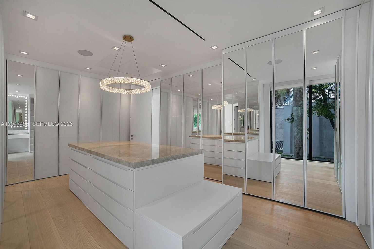 This expansive walk-in closet features a central island with a stone countertop and integrated storage drawers, complemented by light wood flooring. The room is lined with floor-to-ceiling mirrored wardrobe doors that reflect the space, creating an airy and open atmosphere. A modern circular crystal chandelier serves as a focal point, while large windows provide a view of the outdoor greenery.