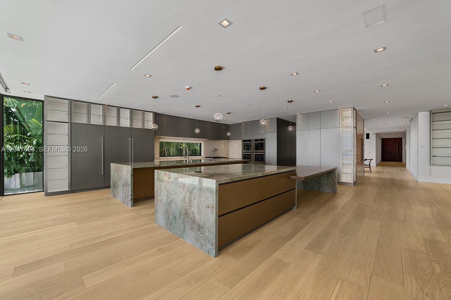 This expansive, modern kitchen features a large central island with a stone countertop and integrated seating, complemented by dark cabinetry and light wood flooring. The space is illuminated by multiple pendant lights and recessed ceiling lighting, creating a bright and open atmosphere. A long hallway extends into the distance, while large windows on the left provide a view of lush greenery outside.