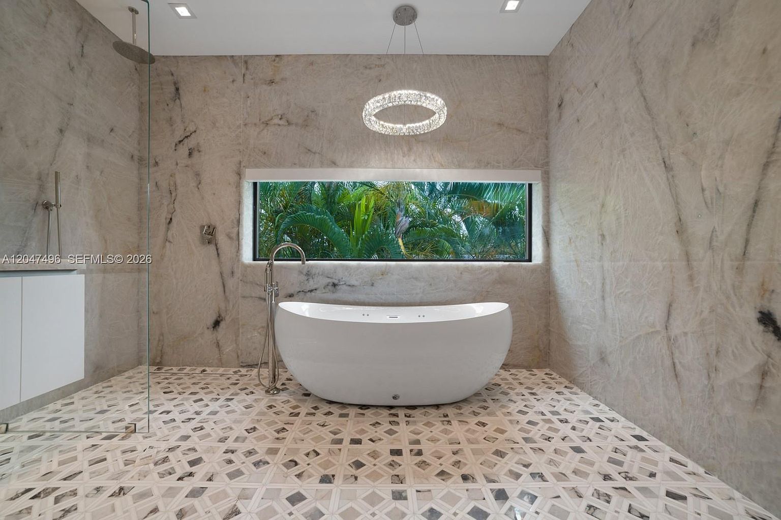 This luxurious primary bathroom features a freestanding soaking tub centered against a wall clad in large-format, veined stone panels. A horizontal window above the tub provides a serene view of lush tropical greenery, while a modern, circular crystal chandelier hangs overhead. The space is finished with intricate, geometric patterned tile flooring and includes a glass-enclosed shower area to the left.