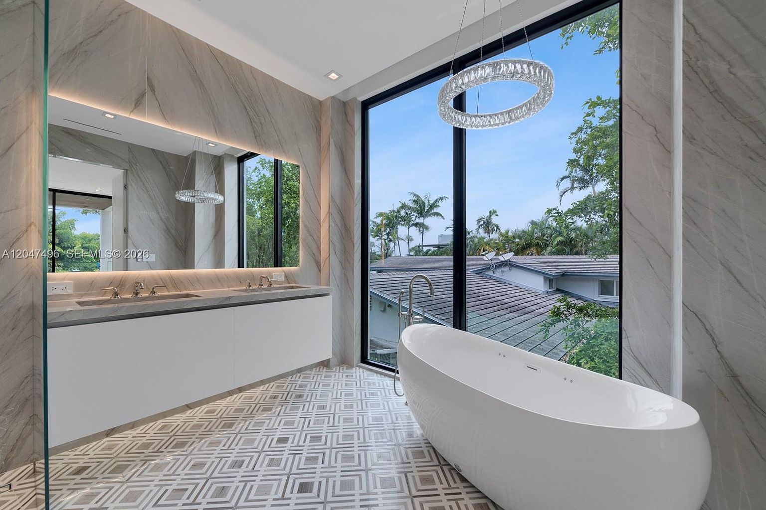 This luxurious primary bathroom features a sleek, wall-mounted double vanity with a large, integrated mirror and stone-clad walls. A freestanding soaking tub is positioned next to expansive floor-to-ceiling windows that offer a scenic view of the surrounding trees and neighborhood rooftops. The space is finished with intricate, geometric patterned floor tiles and a modern circular chandelier, creating a sophisticated and airy atmosphere.