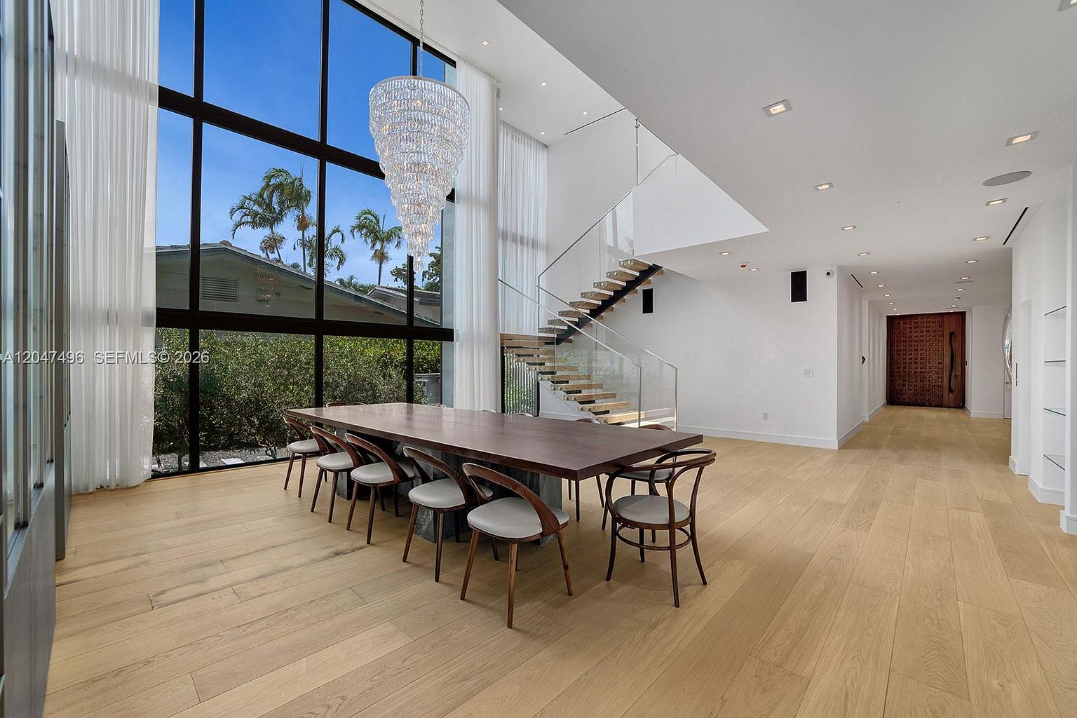 This spacious dining area features a large, dark wood table surrounded by curved-back chairs, set against a backdrop of expansive floor-to-ceiling windows that flood the room with natural light. A dramatic, multi-tiered crystal chandelier hangs above the table, while a modern floating staircase with glass railings adds a sophisticated architectural element to the open-concept space. The room is finished with light wood flooring and clean white walls, creating an airy and luxurious atmosphere.