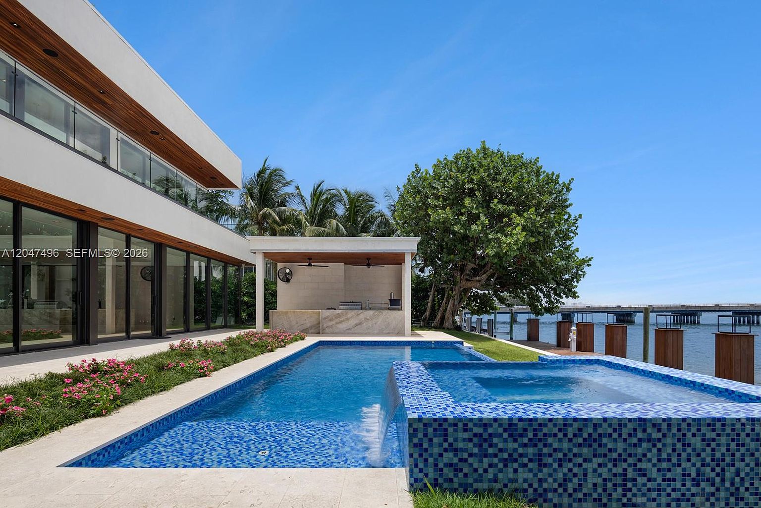 This modern waterfront property features a sleek, rectangular swimming pool with an integrated raised spa that spills over into the main pool. Adjacent to the water, a covered outdoor kitchen area provides a shaded space for entertaining, while the home's contemporary architecture is showcased through large glass panels. The scene is set against a backdrop of a clear blue sky and a distant bridge, emphasizing the luxurious, resort-like atmosphere of the backyard.