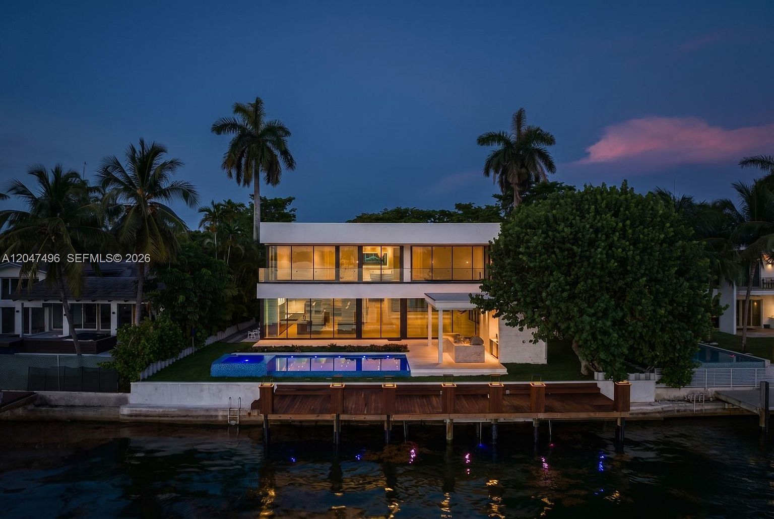 This modern, two-story waterfront residence is captured at dusk, showcasing a sleek architectural design with expansive glass walls that offer a seamless view of the interior. The property features a private swimming pool, a covered outdoor lounge area, and a wooden dock extending into the calm water. Lush tropical landscaping, including tall palm trees, frames the home, creating a sophisticated and serene atmosphere.