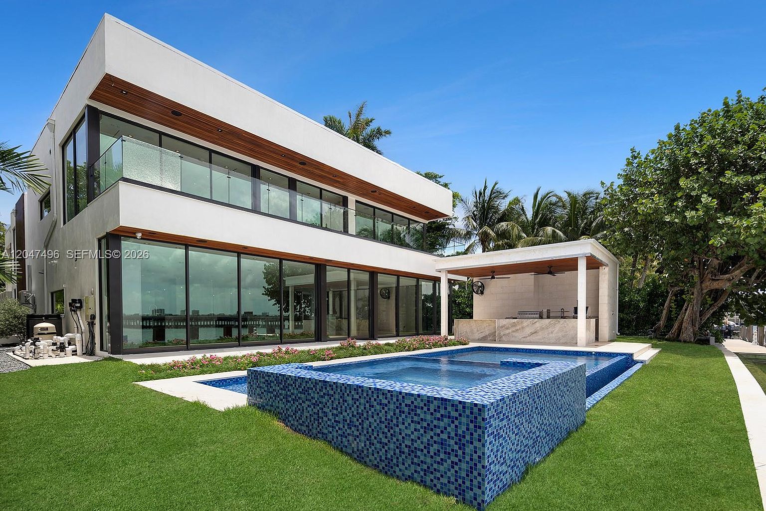 This modern, two-story residence features a sleek white facade with extensive floor-to-ceiling glass walls that provide a seamless connection to the outdoors. The backyard includes a custom-tiled swimming pool with a raised spa section and a covered outdoor kitchen area with a stone-topped bar. Lush green lawn and mature tropical landscaping frame the property, creating a private and luxurious waterfront atmosphere.