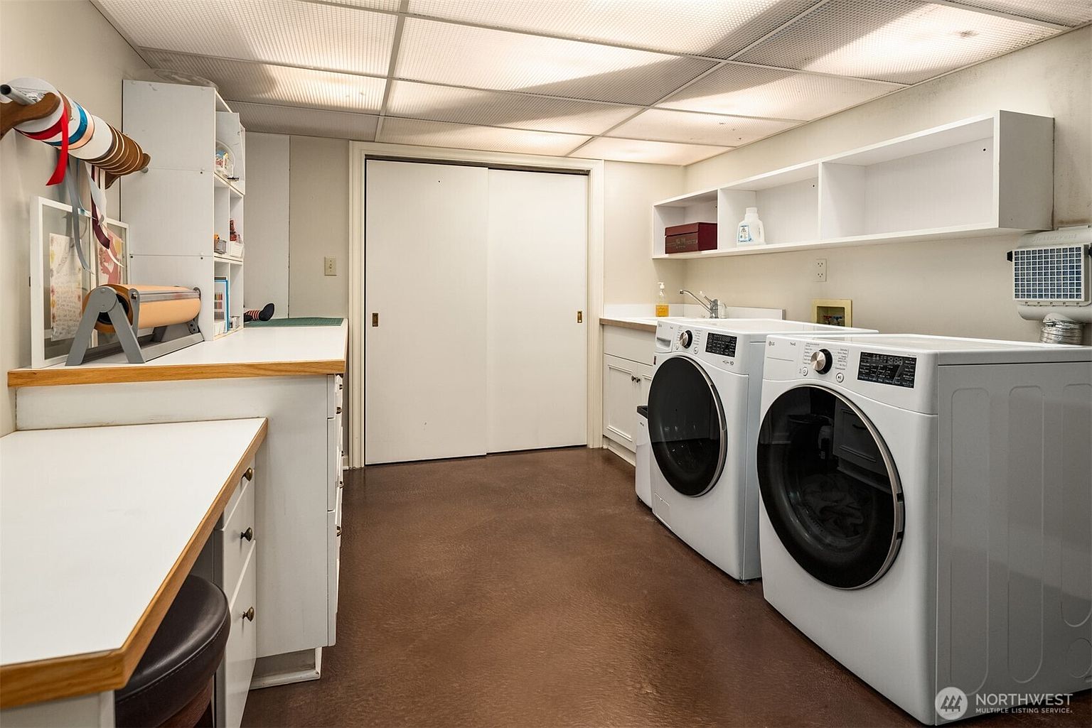 This is a well-organized laundry room featuring a pair of modern, white front-loading washing machines and dryers. The room includes ample counter space with built-in cabinetry and open shelving for storage. The floor is a warm brown, and the lighting is bright, creating a clean and functional space.