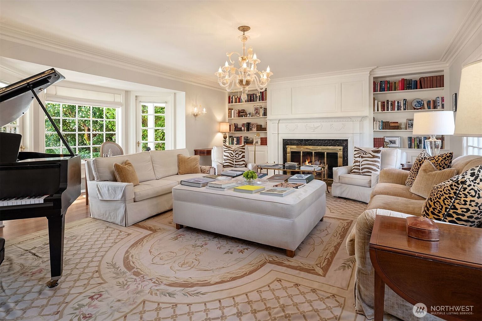 This is a well-lit living room featuring a grand piano, a large sofa, and a fireplace flanked by built-in bookshelves. The room is decorated in a traditional style with a neutral color palette, accented by patterned pillows and a decorative rug. A chandelier hangs from the ceiling, adding a touch of elegance.
