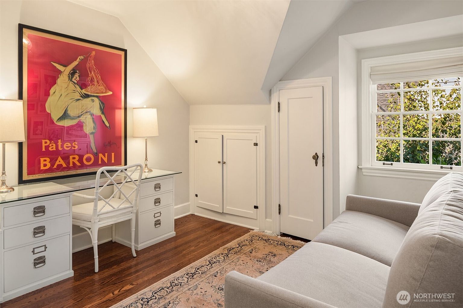 This is an interior shot of a cozy office or study space. The room features a desk with a glass top and white drawers, a white chair, and a large framed poster on the wall. A small sofa sits near a window, and a patterned rug covers part of the hardwood floor, creating a warm and inviting atmosphere.
