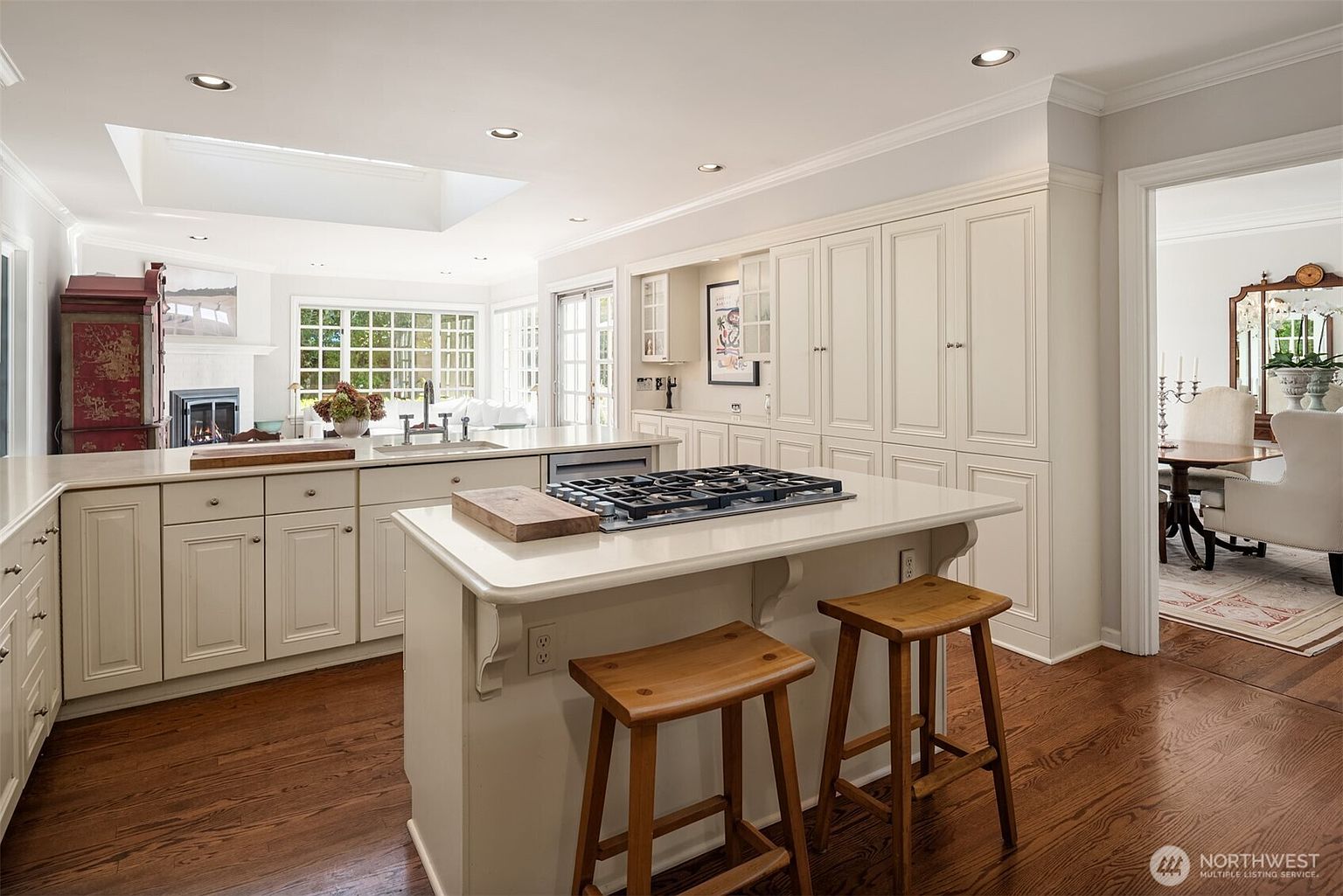 This is a bright and spacious kitchen featuring custom white cabinetry, a large center island with a gas cooktop and seating, and hardwood floors. The kitchen is well-lit with recessed lighting and a skylight, and it opens to a dining area, creating an inviting and functional space. The overall impression is one of elegance and practicality, perfect for both cooking and entertaining.