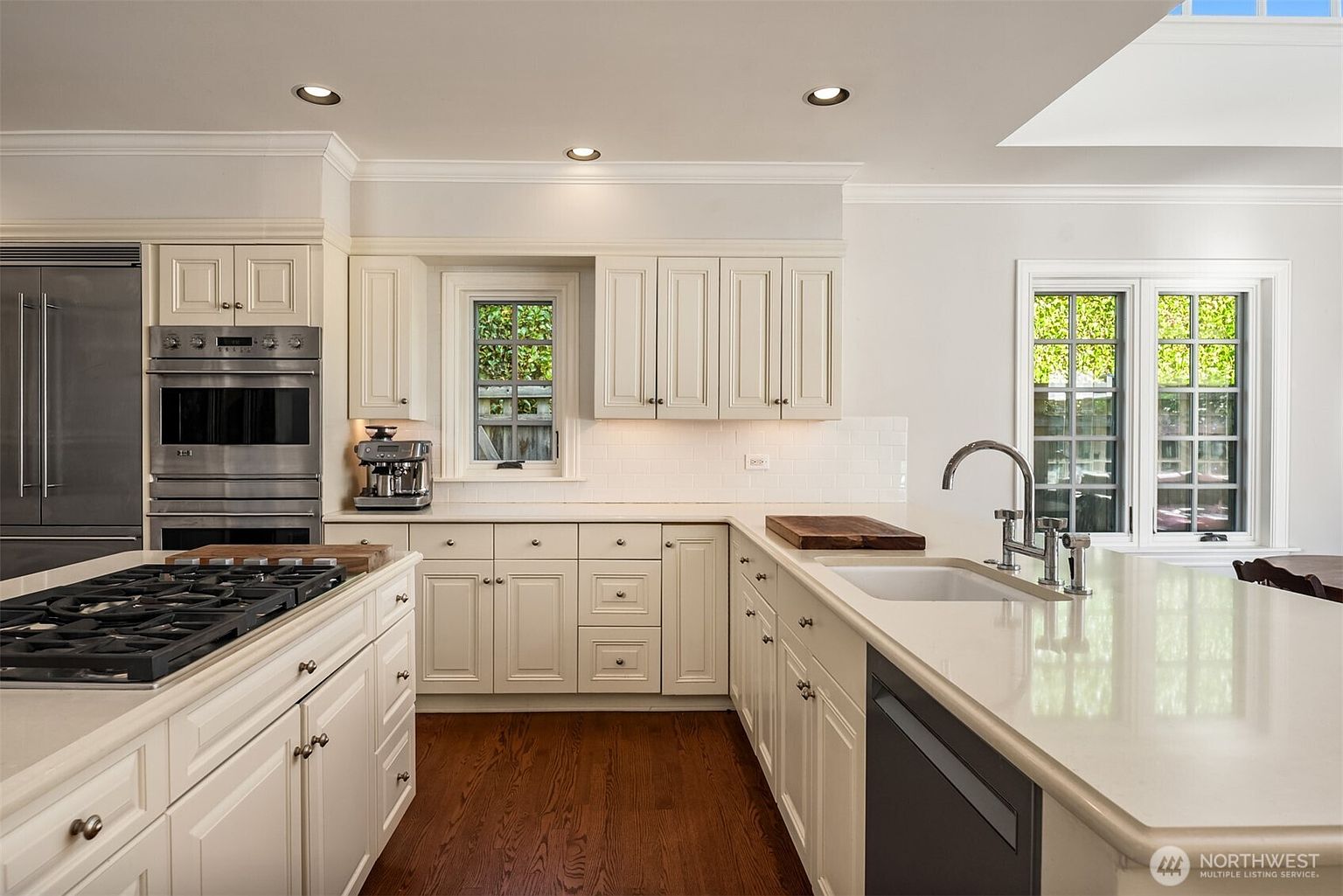 This is a well-lit kitchen featuring custom white cabinetry, stainless steel appliances, and hardwood floors. The kitchen island has a white countertop and a built-in cooktop. Natural light streams in through the windows, creating a bright and inviting atmosphere.