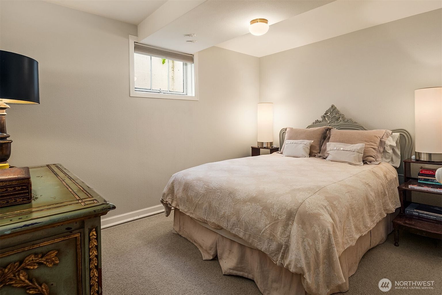 This is a cozy guest bedroom featuring a queen-sized bed with a decorative headboard and neutral-toned bedding. A small window provides natural light, and a bedside table with a lamp adds to the room's ambiance. A dresser with a lamp is also visible, contributing to the room's warm and inviting atmosphere.