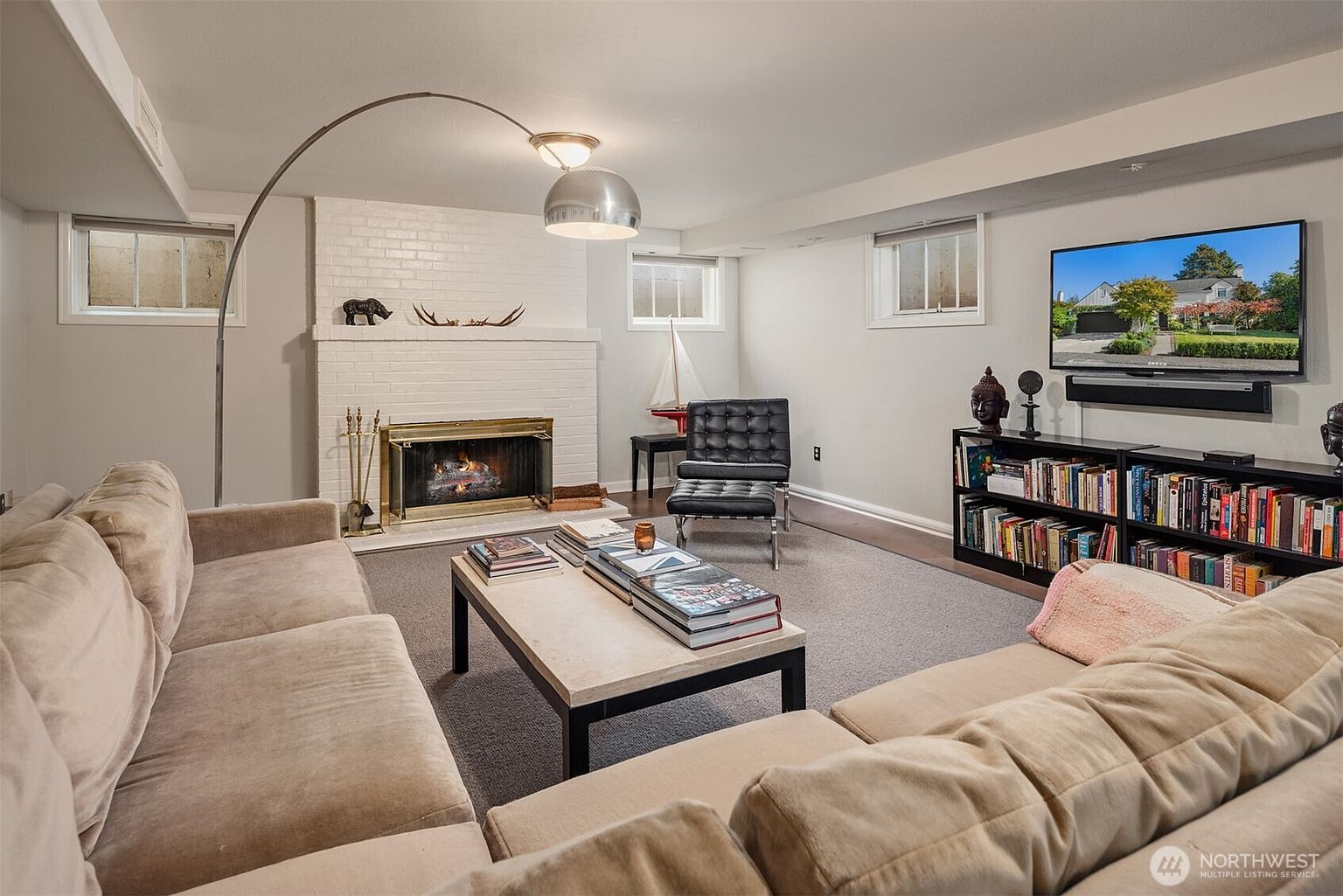 This is an interior shot of a living room featuring a beige sectional sofa, a white brick fireplace with a gold-trimmed firebox, and a modern arc floor lamp. A black bookshelf with a TV mounted above it sits against the far wall, and a coffee table with books is centered in the room. The room has a cozy and inviting atmosphere.