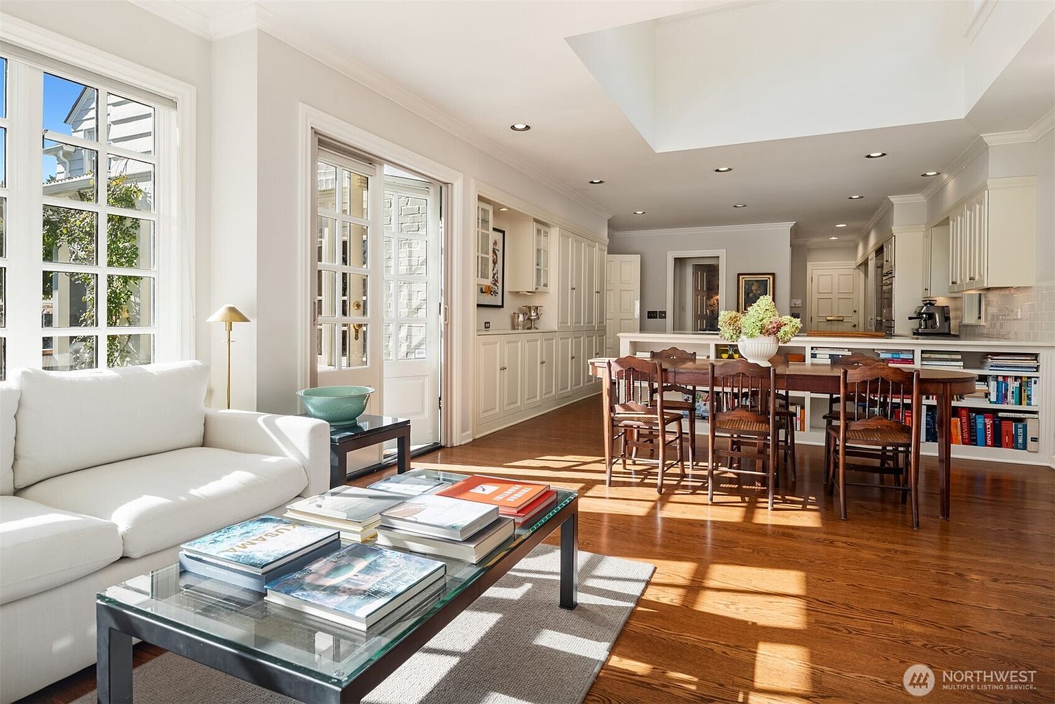 This is an interior shot of a bright and airy living room that seamlessly transitions into a dining area and kitchen. The living room features a comfortable white sofa, a glass-topped coffee table with books, and large windows providing ample natural light. The dining area includes a wooden table with chairs, and the kitchen is equipped with white cabinetry and modern appliances, creating an open and inviting space.