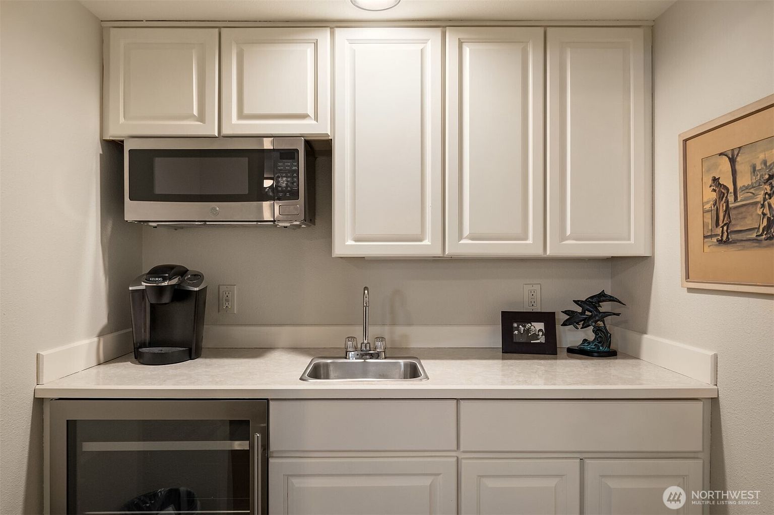 This image showcases a well-organized kitchen area with white cabinetry and a stainless steel microwave and sink. A built-in wine cooler is visible below the counter, and a Keurig coffee maker sits on the countertop. The overall impression is clean and functional, suggesting a modern and efficient space.