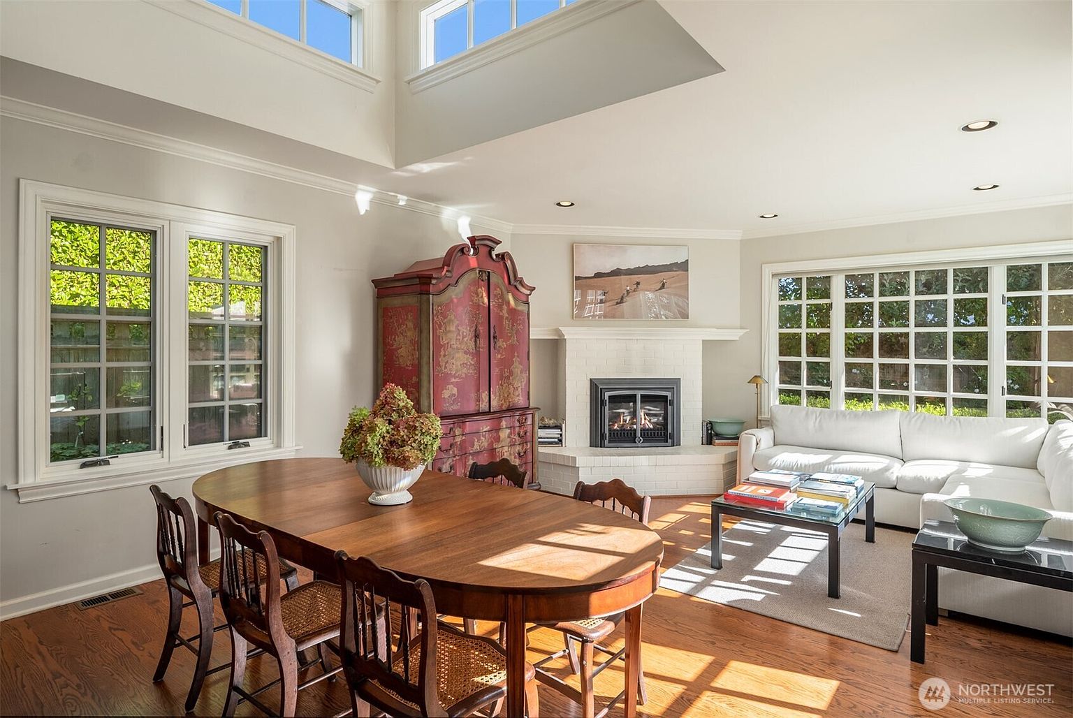 This is an interior shot of a dining room featuring a large wooden dining table with chairs, a red and gold armoire, and a fireplace. Natural light floods the room through large windows, highlighting the hardwood floors and creating a warm, inviting atmosphere. The room also includes a white sofa and coffee table, suggesting a combined living and dining space.