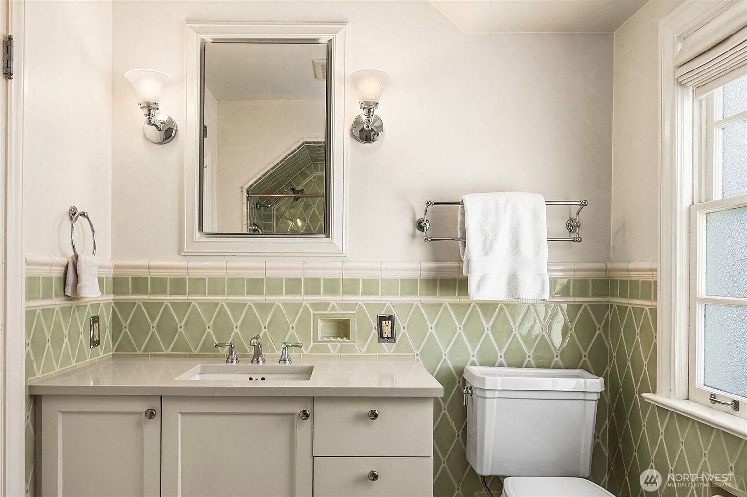 This is a well-lit bathroom featuring a vanity with a light-colored countertop and cabinetry, a sink with chrome fixtures, and a framed mirror flanked by sconce lighting. The walls are painted in a neutral tone, complemented by green and white patterned tiles along the lower portion of the wall. A toilet is visible to the right, and a window provides natural light, creating a clean and inviting space.