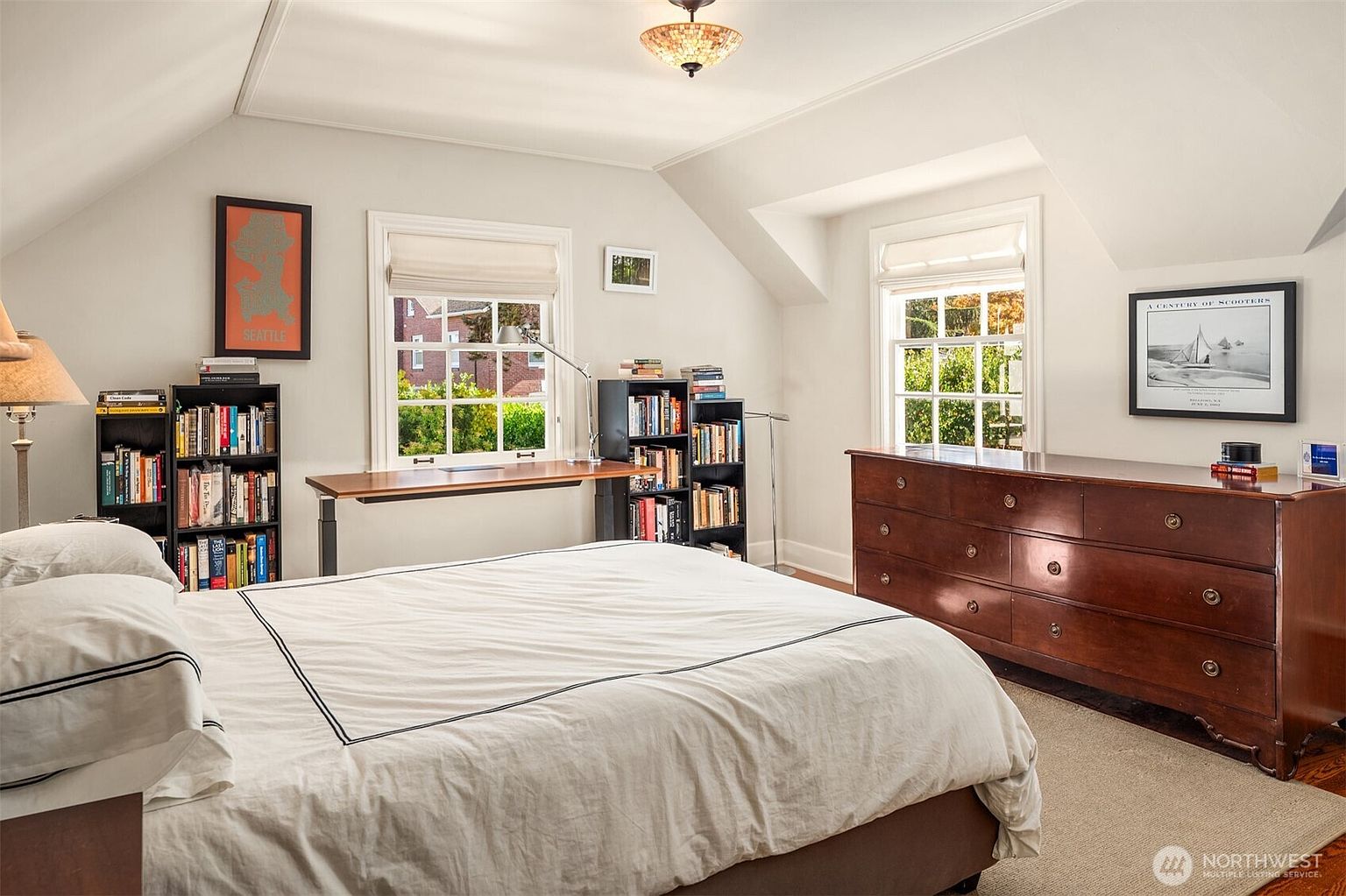 This is a well-lit primary bedroom featuring a large bed with white linens and a dark trim. The room includes a desk area with bookshelves, a dresser with artwork above, and two windows that provide natural light. The overall style is cozy and inviting, suitable for relaxation and rest.