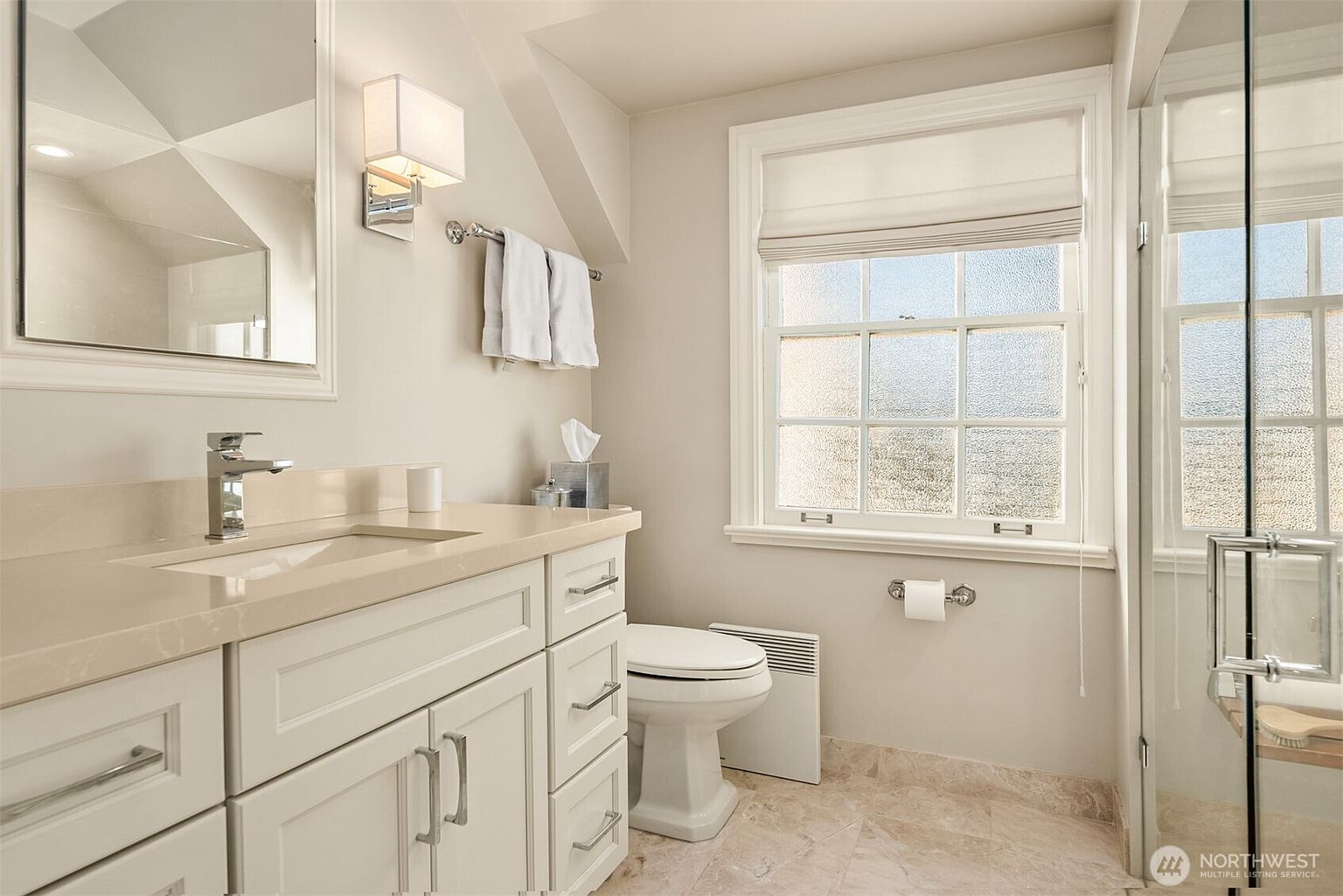 This is a well-lit bathroom featuring a white vanity with a light-colored countertop, a chrome faucet, and ample storage. A framed mirror hangs above the vanity, complemented by modern sconces. The room includes a toilet, a window with a shade, and a glass-enclosed shower, all contributing to a clean and functional space.