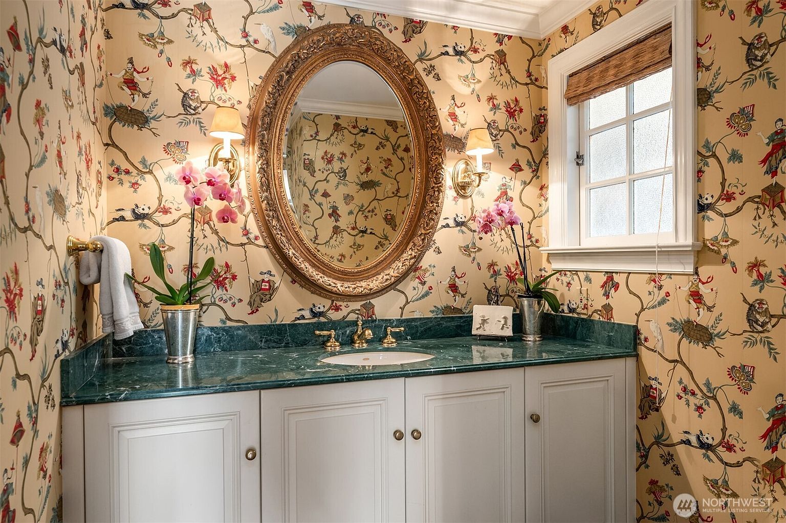 This is a charming guest bathroom featuring a green marble countertop and white cabinetry. The walls are adorned with whimsical wallpaper depicting birds and figures, complemented by an ornate oval mirror and sconce lighting. A window with a woven shade provides natural light, creating a cozy and inviting space.
