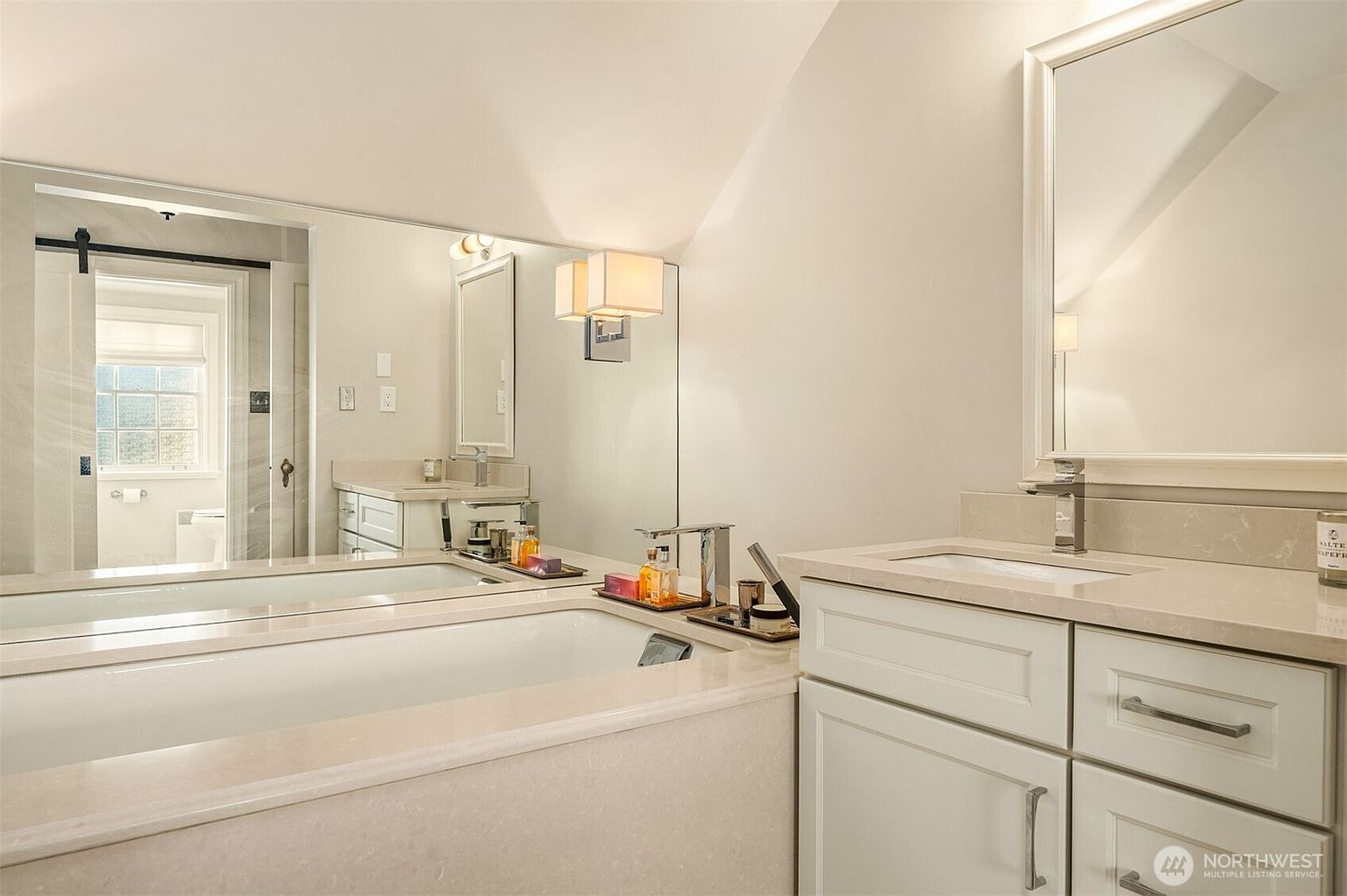 This is a bright and elegant primary bathroom featuring a large soaking tub with a marble surround. The room includes dual vanities with white cabinetry and marble countertops, complemented by modern chrome fixtures. A large mirror reflects the space, enhancing the sense of openness and luxury.