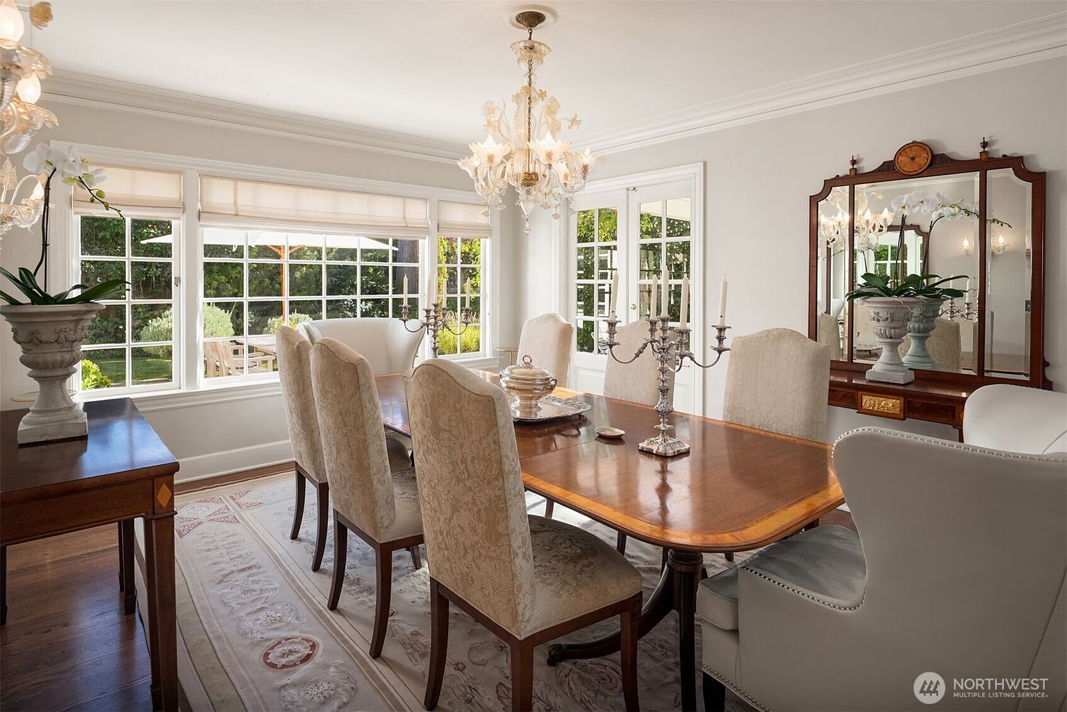 This is an interior shot of a dining room featuring a large wooden table surrounded by upholstered chairs. A crystal chandelier hangs above the table, and a sideboard with a mirror is visible against the wall. The room is well-lit with natural light coming through the windows, creating a bright and inviting atmosphere.