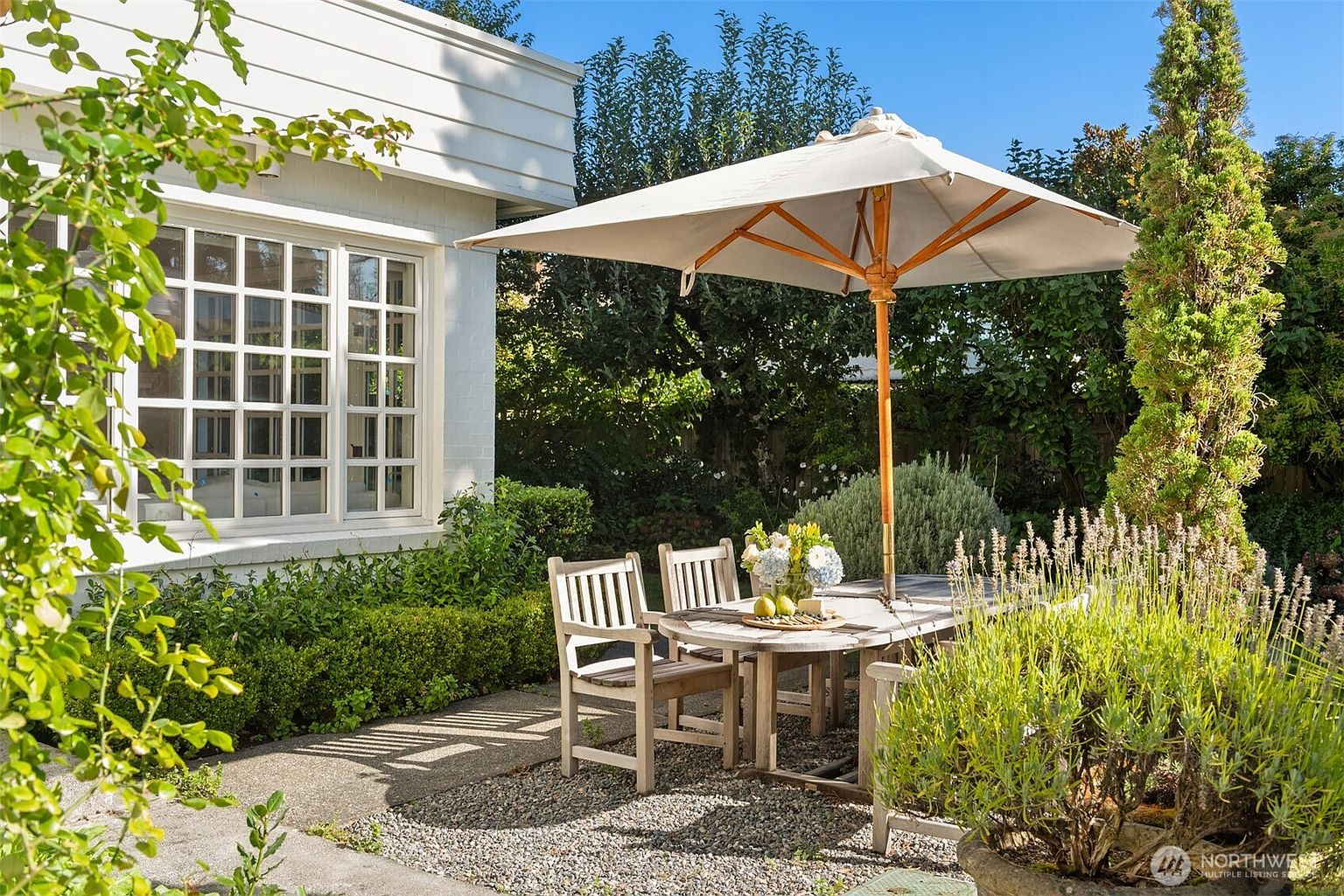 This image showcases a charming outdoor dining area set within a well-maintained garden. A wooden table and chairs are arranged under a large umbrella, creating a shaded space for relaxation and entertaining. The surrounding greenery includes manicured hedges, flowering plants, and a mature tree, contributing to a serene and inviting atmosphere.