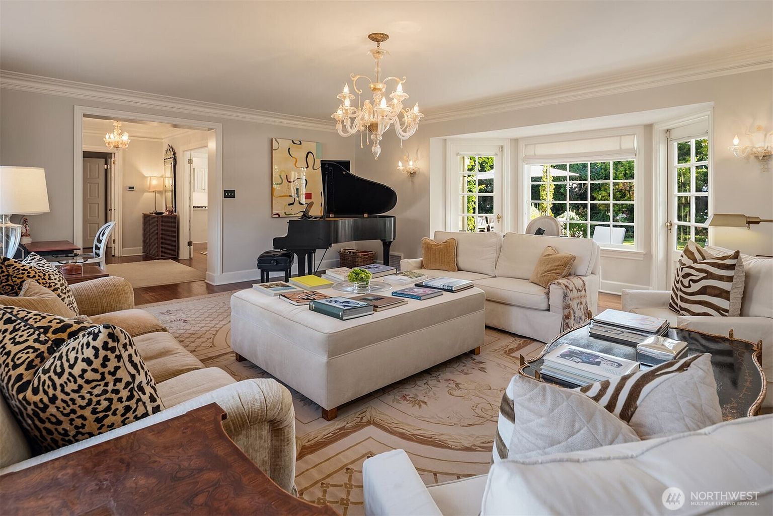 This is a well-lit living room featuring a grand piano, a large ottoman covered with books, and multiple sofas and armchairs arranged around a patterned rug. The room has a classic and elegant style, with a chandelier and large windows providing ample natural light. The space is designed for comfortable living and entertaining.
