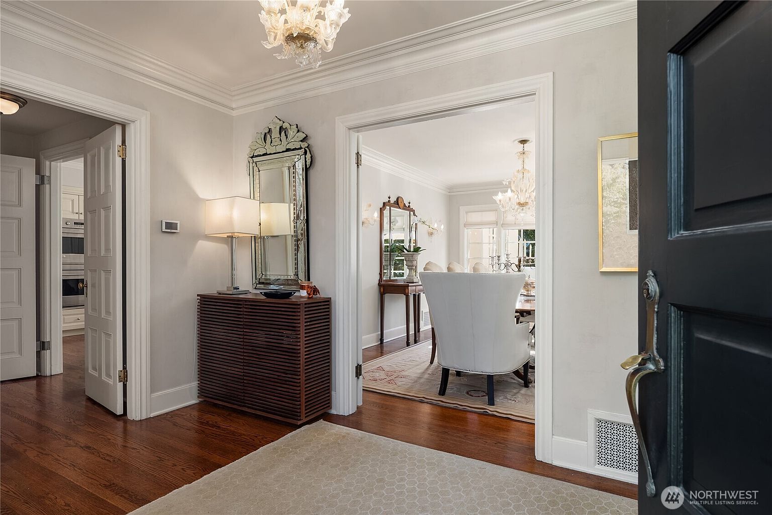 This interior shot showcases an elegant hallway leading into a dining area. The hallway features dark hardwood floors, light gray walls, and sophisticated decor, including a decorative mirror and a stylish cabinet with a lamp. The open doorway provides a glimpse into the dining room, highlighting a chandelier and a formal dining set, creating an inviting and luxurious atmosphere.