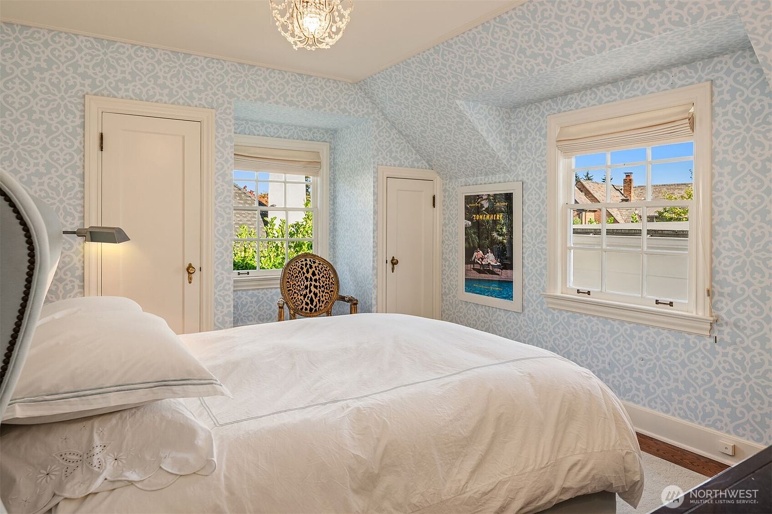 This is a charming bedroom featuring a bed with white linens, light blue patterned wallpaper, and natural light streaming through two windows. A decorative chair and a framed poster add character to the room. The overall impression is cozy and inviting, perfect for a guest room.