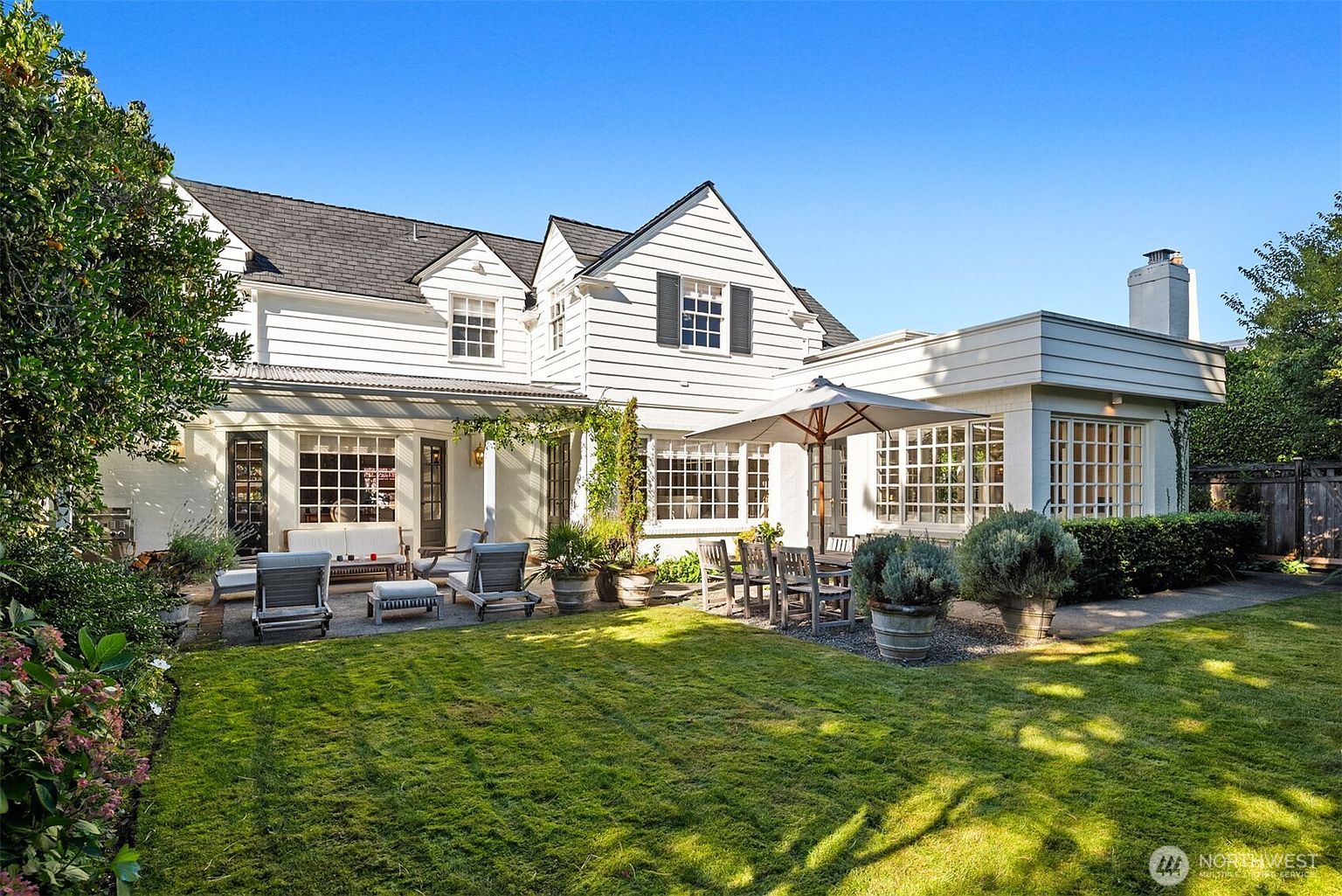 This is a rear view of a charming two-story house with a well-manicured lawn and patio area. The house features white siding, black shutters, and a dark roof. The patio is furnished with outdoor seating and a dining set, perfect for entertaining or relaxing. The overall impression is one of a well-maintained and inviting outdoor space.