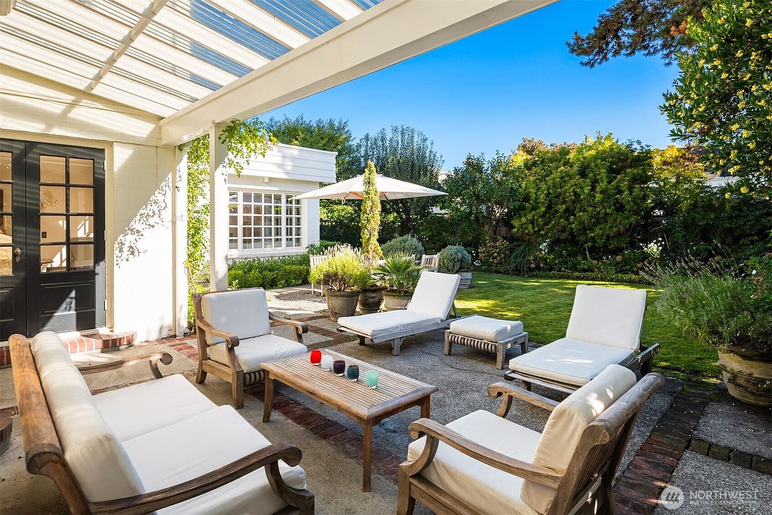This image showcases a beautifully arranged outdoor patio area, perfect for relaxation and entertaining. The space features comfortable seating arrangements with cushioned chairs and a chaise lounge, all centered around a wooden coffee table. A pergola provides partial shade, while the surrounding lush garden and manicured lawn create a serene and inviting atmosphere.