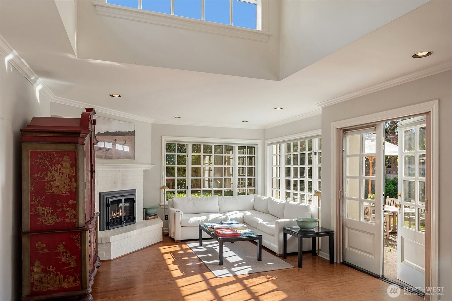 This is an interior shot of a living room featuring a white sectional sofa, a fireplace with a white tile surround, and large windows that provide ample natural light. The room has hardwood floors and a neutral color palette, creating a bright and airy atmosphere. A decorative cabinet adds a touch of color and character to the space.