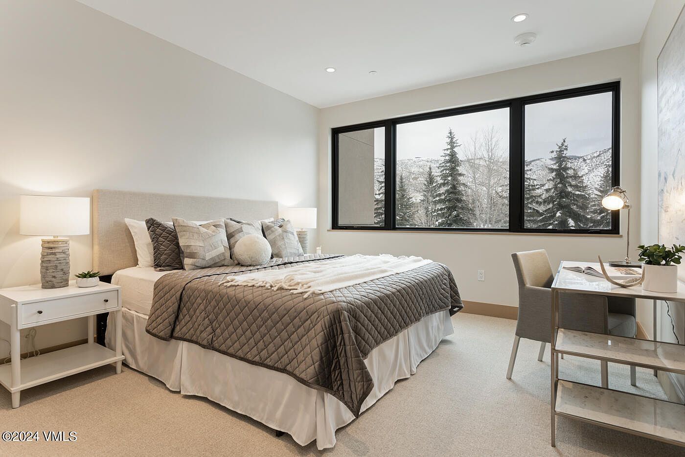 This serene bedroom features a neutral color palette with a plush, quilted bed as the focal point, complemented by a modern desk area. Large windows frame a picturesque, snow-covered forest view, bringing natural light and a tranquil atmosphere into the space. The room is styled with contemporary furnishings, including a bedside table with a textured lamp and a sleek desk, creating a sophisticated and inviting retreat.
