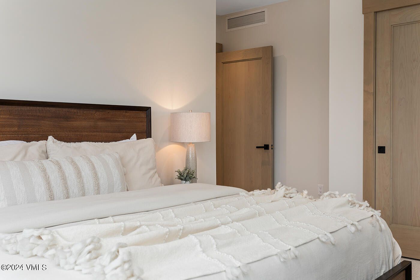 This serene bedroom features a neutral color palette with a textured, cream-colored throw blanket draped over the bed, complemented by a dark wood headboard. A stylish table lamp sits on a nightstand, casting a warm, inviting glow against the soft beige walls. The room includes light wood doors with modern black hardware, creating a clean, contemporary aesthetic that feels both cozy and sophisticated.