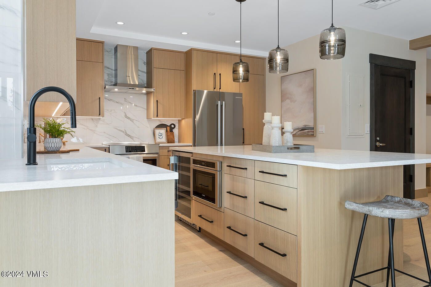 This modern, high-end kitchen features light wood cabinetry, sleek stainless steel appliances, and a spacious island topped with white quartz. Three stylish pendant lights hang above the island, which includes a built-in microwave and wine cooler, while a marble-patterned backsplash adds a touch of elegance. The perspective is a wide-angle shot from the kitchen island, showcasing a clean, contemporary design perfect for entertaining.