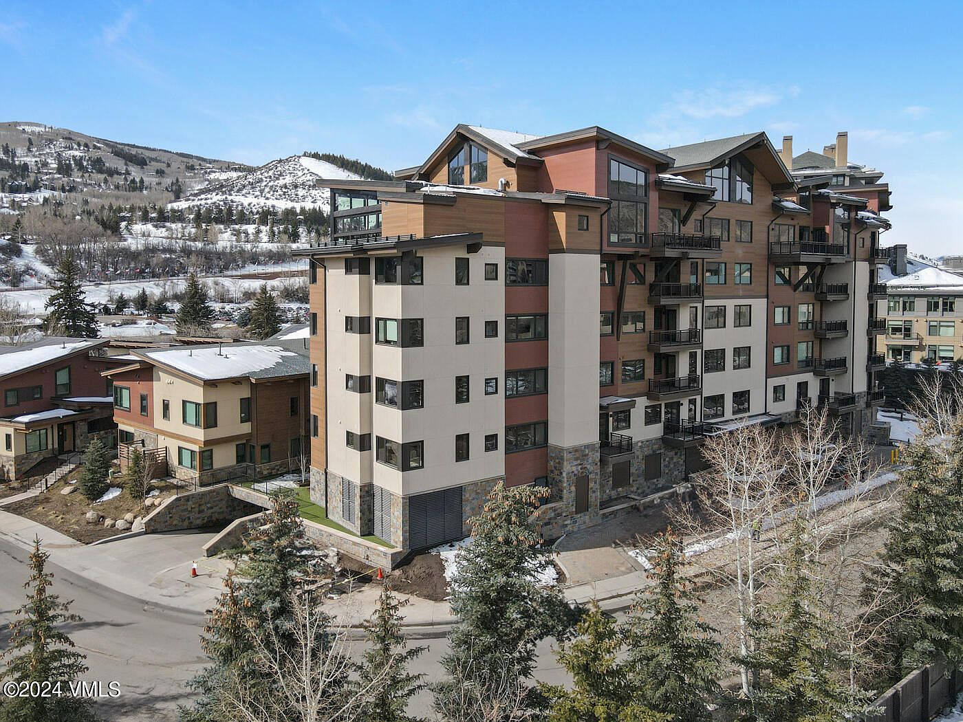 This aerial perspective captures a multi-story, modern mountain-style condominium complex featuring a blend of stone, stucco, and wood siding. The building is situated in a snowy, alpine environment with clear blue skies and surrounding evergreen trees. The architecture emphasizes a contemporary aesthetic with tiered balconies and large windows, reflecting a high-end residential development.