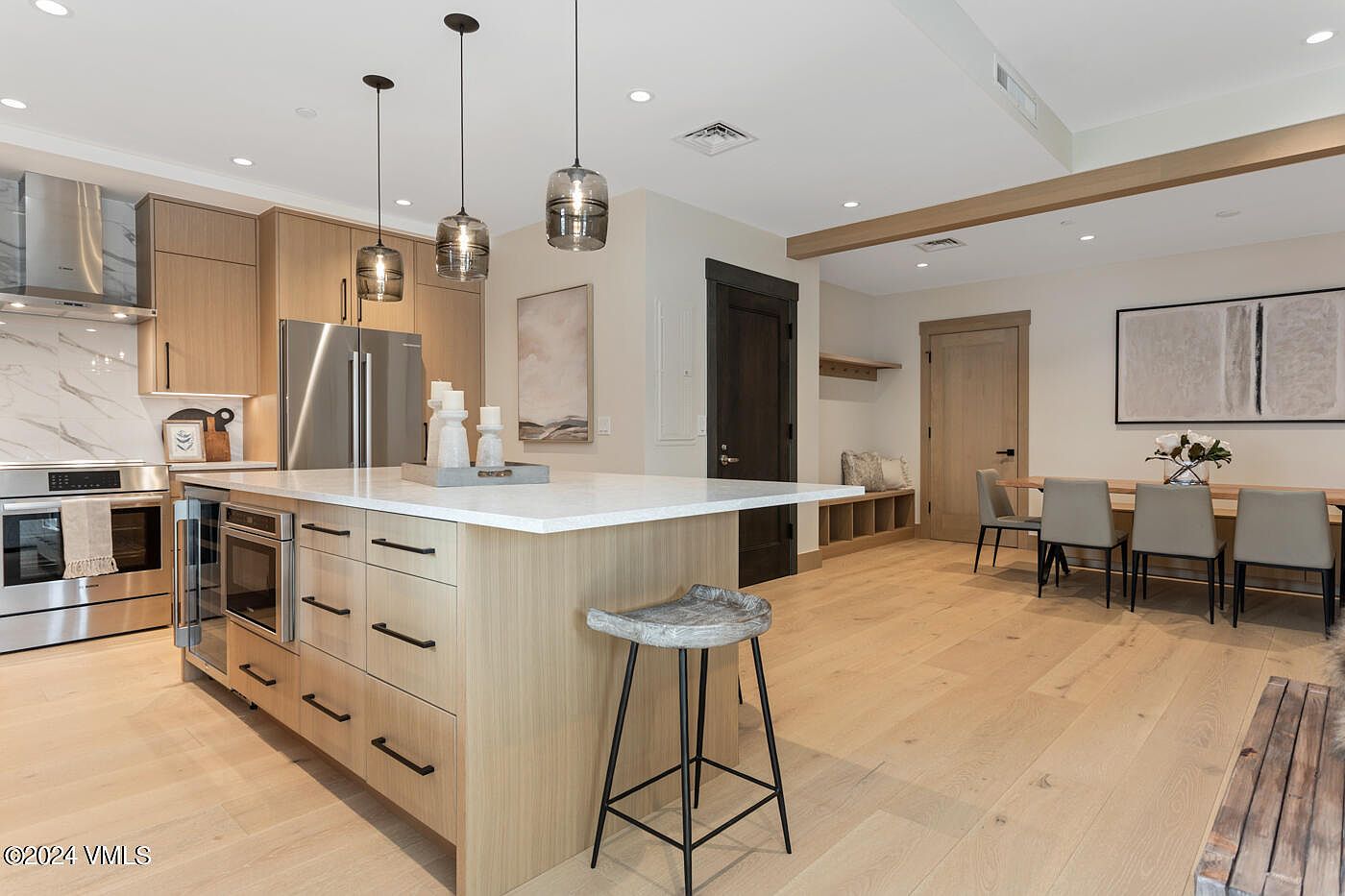 This modern, open-concept kitchen features light wood cabinetry, a large central island with a white quartz countertop, and high-end stainless steel appliances. Three glass pendant lights hang above the island, while the adjacent dining area and built-in mudroom bench create a seamless, functional living space. The light hardwood flooring and neutral color palette contribute to a bright, airy, and contemporary aesthetic.