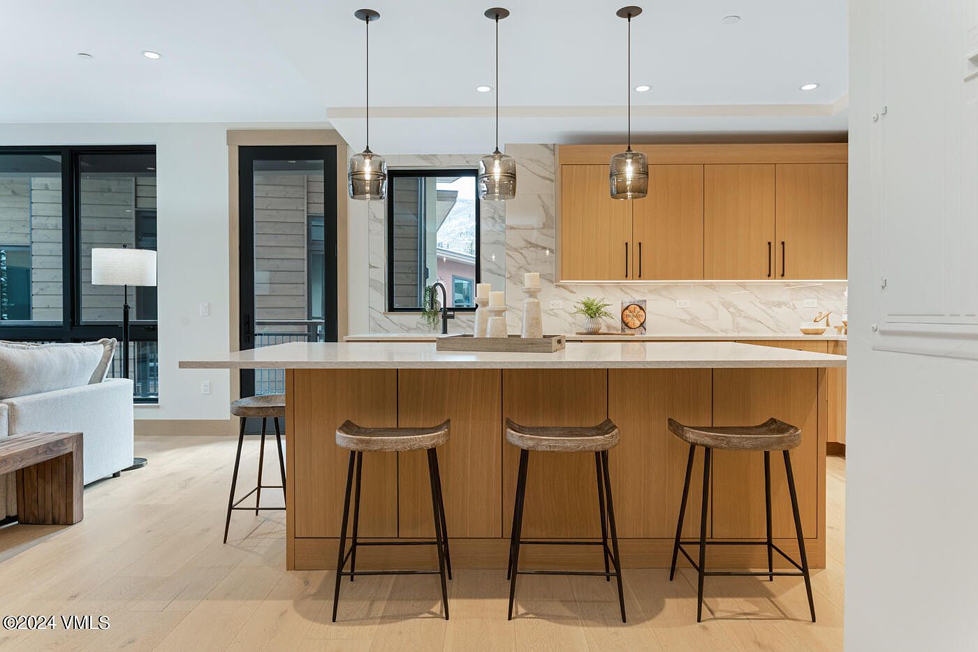This modern, open-concept kitchen features light wood cabinetry, a large central island with a stone countertop, and three rustic bar stools. Three glass pendant lights hang above the island, complementing the sleek marble backsplash and contemporary aesthetic. The perspective is a straight-on, eye-level shot that highlights the seamless flow between the kitchen and the adjacent living area.