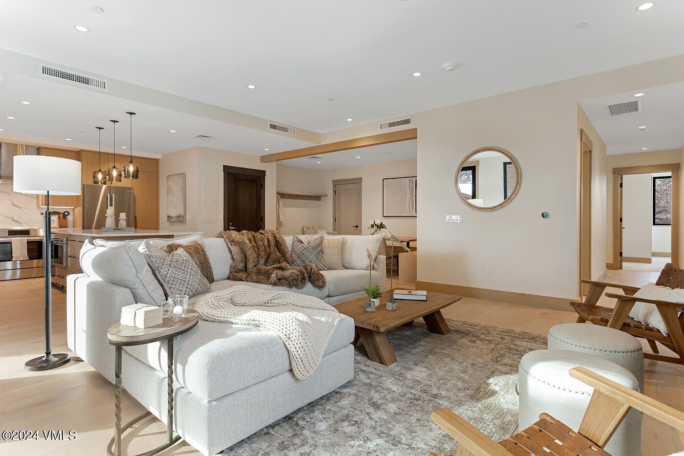 This bright and inviting living room features a large, plush sectional sofa adorned with textured throw pillows and a cozy blanket, centered around a rustic wooden coffee table. The open-concept layout seamlessly connects to a modern kitchen in the background, while a circular wall mirror and neutral color palette enhance the sense of space and light. The perspective is from a low-angle, wide-shot view that captures the comfortable, high-end residential atmosphere.
