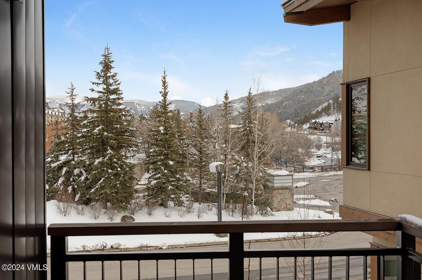 This image captures a scenic view from a balcony overlooking a snow-covered landscape with tall pine trees and distant mountains. The perspective is from an elevated position, showcasing a modern building's exterior railing and siding in the foreground. The overall atmosphere is serene and wintry, highlighting the property's picturesque mountain location.