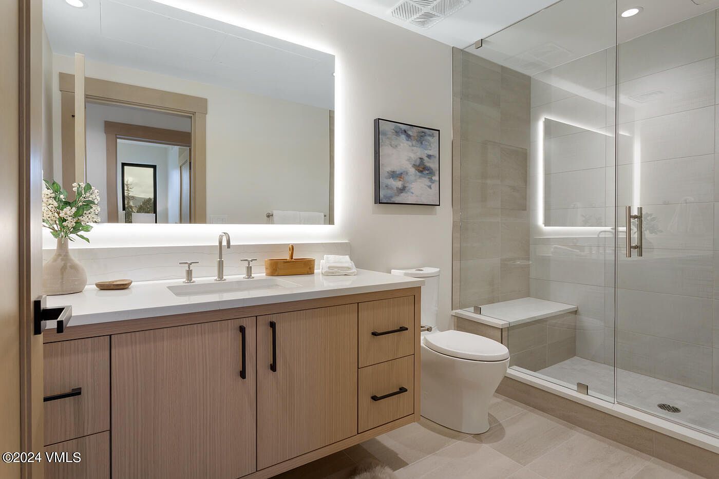 This modern primary bathroom features a sleek floating vanity with light wood cabinetry, a white quartz countertop, and a large, illuminated mirror. A spacious walk-in glass shower with neutral-toned tile and a built-in bench adds a luxurious, spa-like feel to the room. The overall aesthetic is clean, contemporary, and bright, emphasizing high-end finishes and functional design.