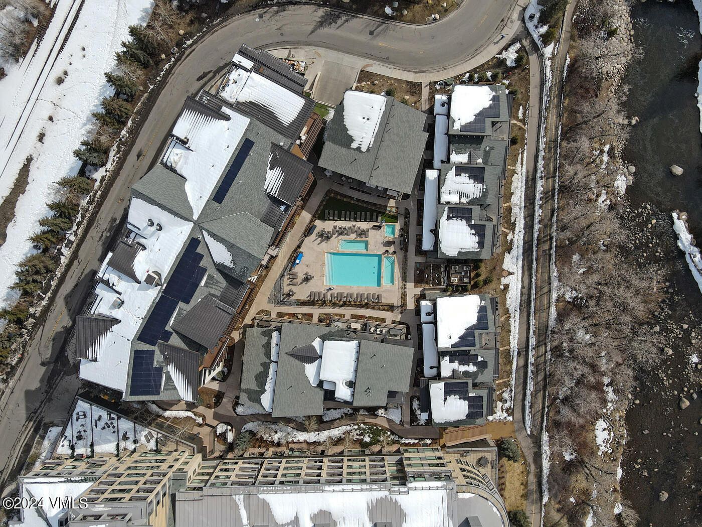 This high-angle aerial view captures a multi-building residential complex nestled in a snowy, mountainous landscape. The property features a central outdoor swimming pool and hot tub area, surrounded by buildings with dark shingled roofs partially covered in snow. A winding road borders the complex on one side, while a river flows along the other, creating a scenic and tranquil setting.