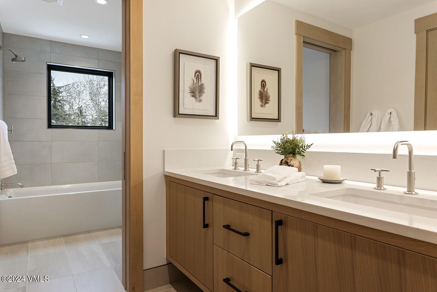 This modern primary bathroom features a sleek double vanity with light wood cabinetry and a white quartz countertop, complemented by minimalist brushed nickel fixtures. A large, illuminated mirror spans the wall above the vanity, while an open doorway provides a view into a separate area containing a bathtub and tiled shower. The space is bright and airy, showcasing a contemporary design with clean lines and a neutral color palette.