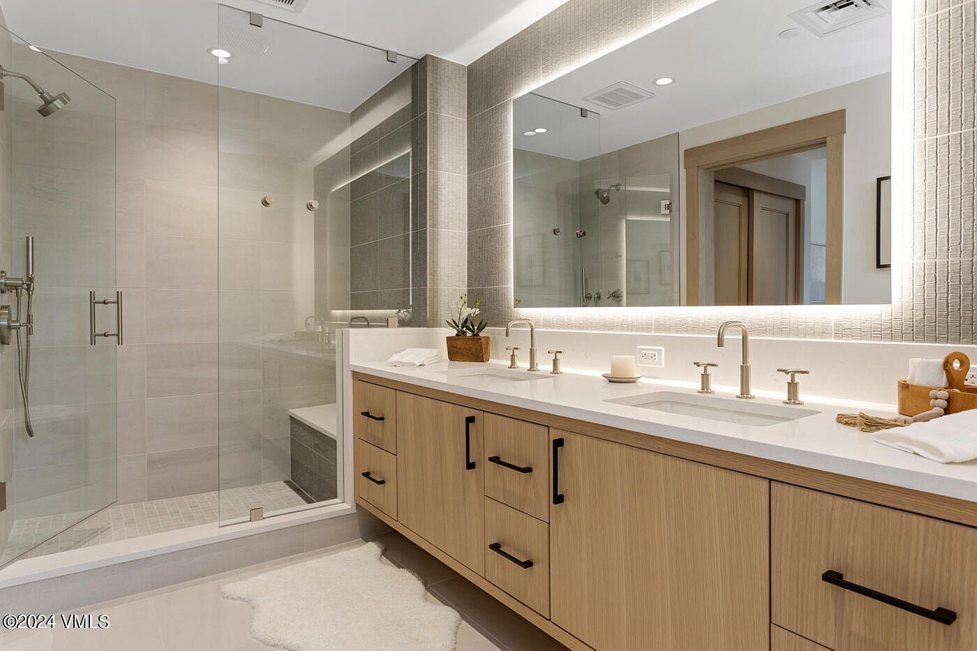 This modern primary bathroom features a sleek double vanity with light wood cabinetry, white quartz countertops, and contemporary matte black hardware. A large, illuminated mirror spans the wall above the sinks, while a spacious walk-in glass shower with neutral tile work sits to the left. The overall aesthetic is clean, bright, and luxurious, offering a spa-like atmosphere.