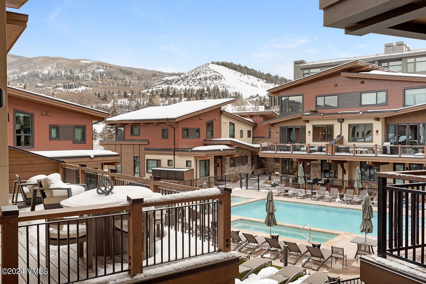 This elevated perspective captures a resort-style outdoor pool and hot tub area nestled within a modern mountain community. The scene features a foreground deck with patio furniture, overlooking the pool deck lined with lounge chairs, all set against a backdrop of snow-dusted residential buildings and a scenic, snow-covered mountain range. The architecture reflects a contemporary alpine aesthetic, emphasizing a luxurious and relaxing atmosphere perfect for year-round mountain living.