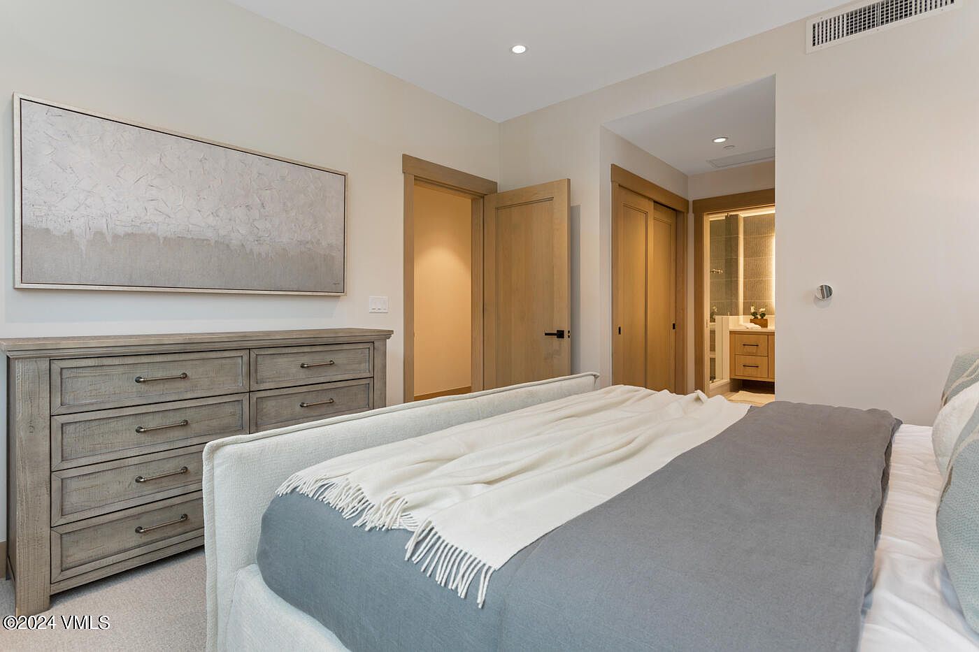 This bedroom features a neutral color palette with a light-toned wooden dresser and matching door frames, creating a warm and inviting atmosphere. A plush bed with a grey and white throw blanket serves as the focal point, while an open doorway leads to a well-lit bathroom. The room is accented by a large, textured abstract painting above the dresser, emphasizing a clean, modern aesthetic.